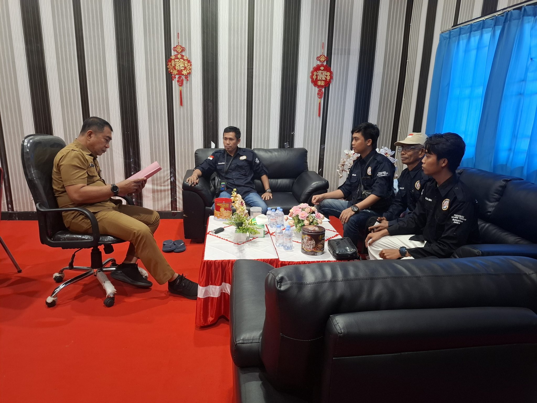 Pejabat berseragam cokelat membaca dokumen di hadapan anggota tim yang duduk dalam ruang pertemuan