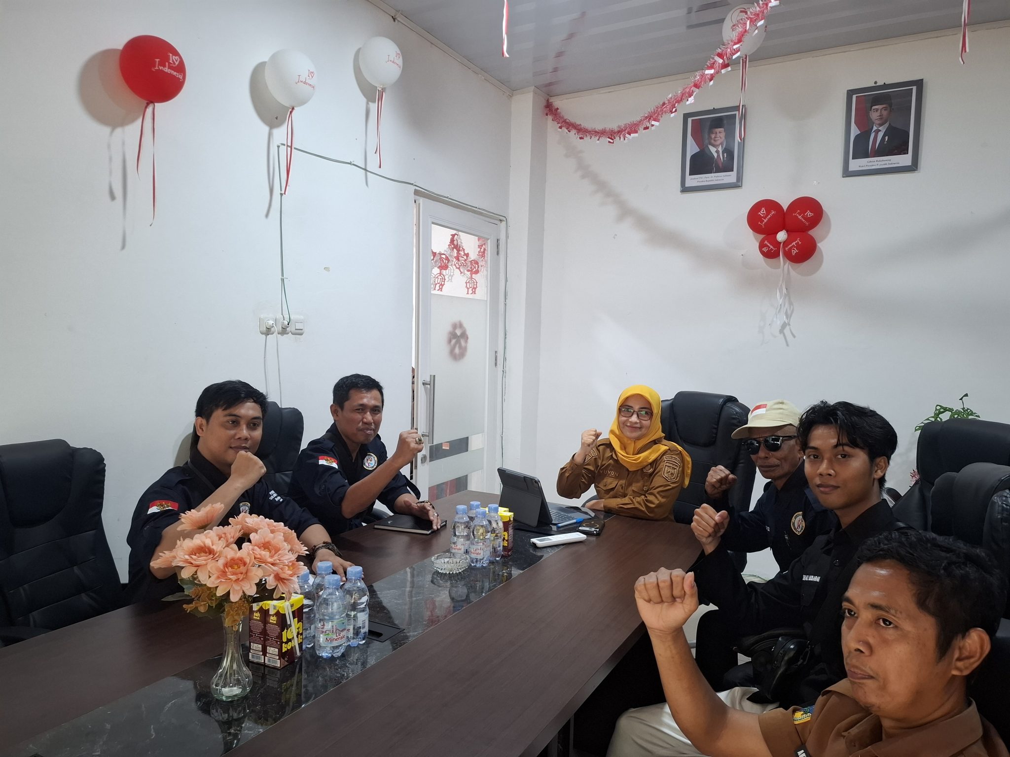 Peserta pertemuan duduk mengelilingi meja, termasuk seorang perempuan berkerudung kuning dan anggota tim berpakaian gelap