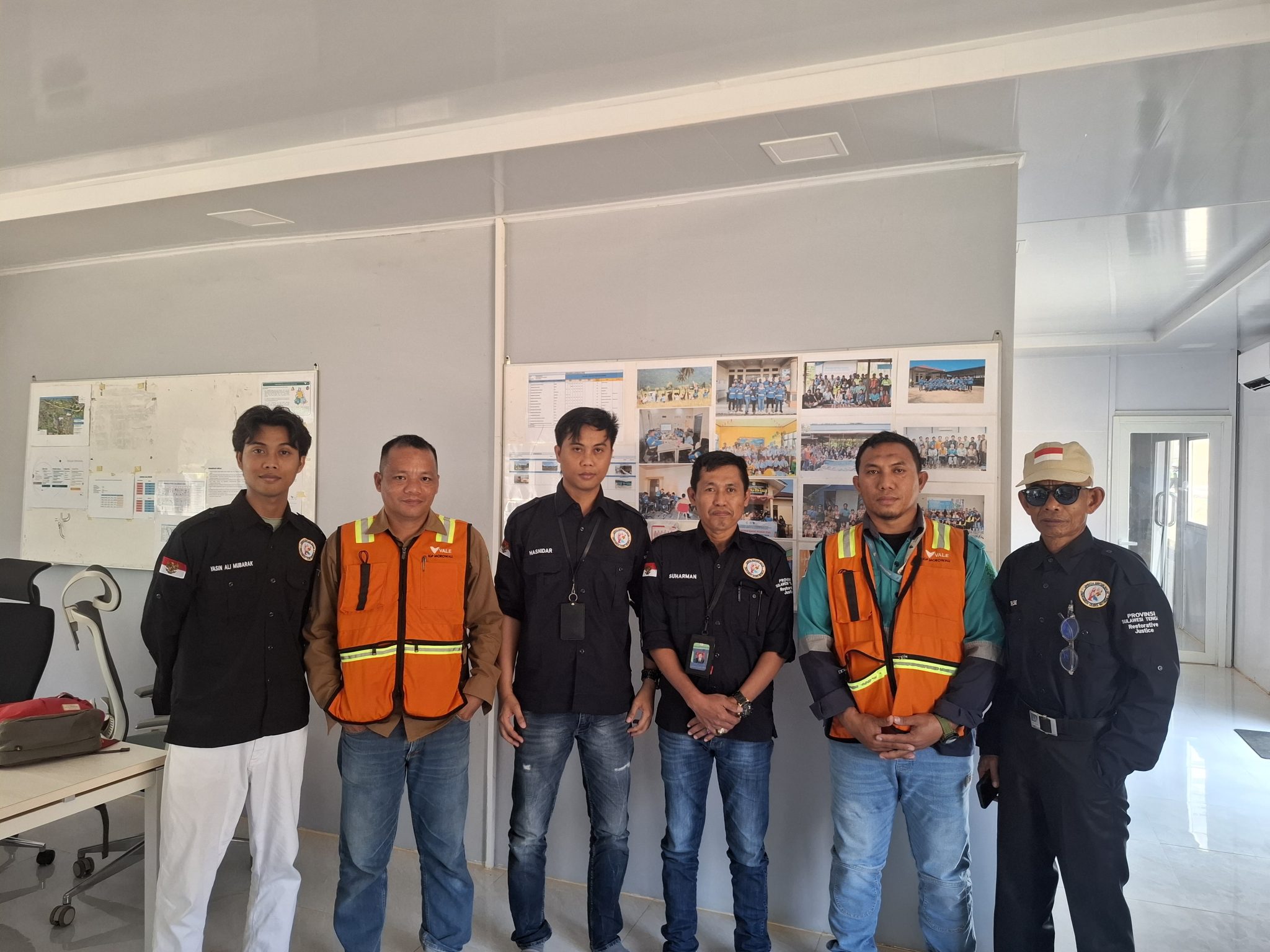 Sekelompok orang berdiri berfoto bersama di dalam ruangan, beberapa mengenakan rompi oranye dan lainnya berpakaian gelap